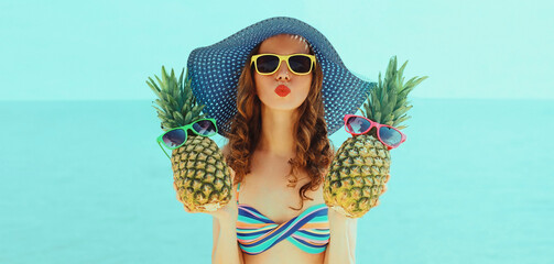 Portrait of young woman blowing her lips sending air kiss with two funny pineapples with sunglasses on the beach wearing straw hat