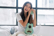 © kaew6566 - Cheerful asian woman hold ukulele sitting on white bed with pillow beside window at apartment. Joyful young girl holding acoustic ukulele in morning at home. Relax female play music with ukulele hobby