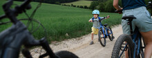 Portrait Of Excited Little Boy With His Family At Backround Riding Bike On Path In Park In Summer