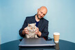 © mark_gusev - Man holing British short hair cat in his hands. Bald male in his 40s, sitting at a table with computer and cup of coffee. Working at home theme. Human annoy busy cat