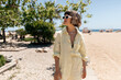 © PhotoBook - Cute stylish european young woman genuinely laughs, relaxing on beach. Short-haired girl in glasses and summer suit walking around on the beach