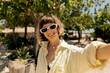 © PhotoBook - Wonderful stylish smiling girl with wavy short hair wearing sunglasses is making selfie in summer sunny day on background of green plants on the beach