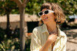 © PhotoBook - Close up portrait of pretty girl with sort wavy hair wearing stylish white sunglasses posing wit lovely smile and looking aside on green plants background
