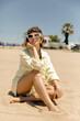 © PhotoBook - Caucasian young tanned girl is sitting on sandy beach in summer outfit. Blonde with short hair looks back smiling with teeth. Healthy lifestyle people concept.
