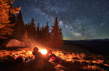 Female Hiker Lying At Karemat Near Bonfire Admire Beauty Of Stunning Starry Sky During Night In Campsite, Backpacker Visiting Mountains With Tent Near Spruce Forest In National Park