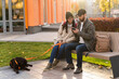 © NFstock - Full length view of happy couple sitting on the street bench at sunset and looking at the screen of device with happy smiles while using smartphone during the walking with the dog