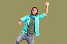 Cheerful Funny Indian Man In Headphones Around His Neck Dancing And Having Fun On Khaki Background. Young Smiling Ethnic Guy In Casual Clothes Having Fun In His Free Time. People Lifestyle Concept.