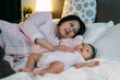 © PR Image Factory - selective focus asian first time mom lying on bed in pajamas is looking at new baby lovingly while putting it to sleep by patting on its chest during nighttime