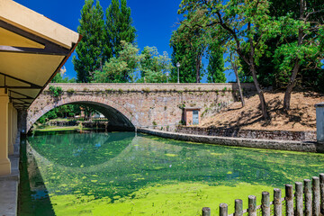 Wall Mural - The ancient wash-house and the masonry bridge over the river, in the medieval village of Bevagna. Perugia, Umbria. Blue sky in a sunny summer day. Green algae on the surface of the stagnant water.