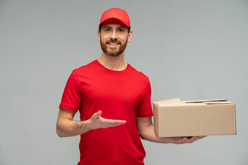 Waist up view portrait of male courier in red uniform handing cardboard parcel to client, carrying parcel with goods ordered online. Indoor studio shot isolated on grey background