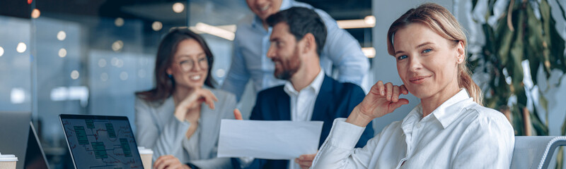 Wall Mural - Smiling confident businesswoman sitting on meeting in office with her colleagues at background.