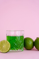 Wall Mural - green alcohol drink in glass near fresh limes on pink background.