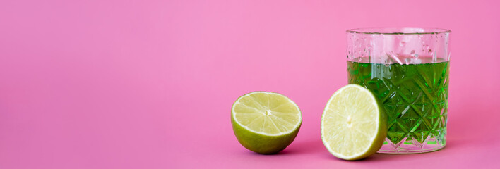 Wall Mural - green alcohol drink in faceted glass with water drops near limes on pink, banner.