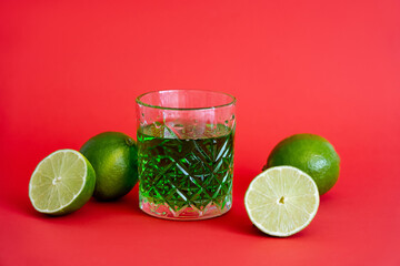 Wall Mural - green alcohol drink in faceted glass near sour limes on red.