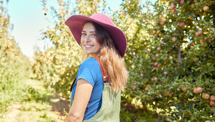 Wall Mural - Portrait of a beautiful female farm worker standing on a fruit farm during harvest season. Young happy farmer between fruit trees on a sunny day in summer. Agricultural industry growing fresh produce