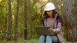 © Yuliia - Attractive woman forestry engineer in protective helmet enters data into laptop takes reforestation action young experienced female specialist ecologist technician watching nature reserve checks trees