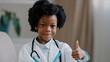 © Yuliia - Close-up little cute kid girl in medical gown looking at camera posing indoors smiling pretending be doctor plays nurse showing thumb up gesture approval promotes dream job future profession concept