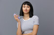 © Tatiana - an attractive, beautiful woman with short black hair stands on a gray background in a striped cotton T-shirt and holds a smartphone in her hand looking at the camera.