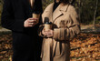 Young man in dark coat and woman in trendy beige coat with cup of coffee outdoor. Beautiful young couple drinks coffee in the autumn park with falling foliage. 