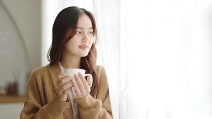 Wall Mural - Young asian woman standing beside window and holding mug in living room at home. Asia female drinking milk or tea after wake up in the morning