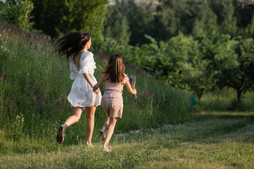 Wall Mural - back view of mother and child holding hands and running on grassy path towards forest.