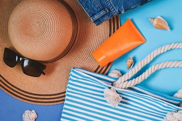 Wall Mural - Summer travel 2022 lifestyle photography. Straw hat, sunglasses, striped bag, orange sunscreen tube and seashells on a blue background. Sea vacation, holidays concept, object photo