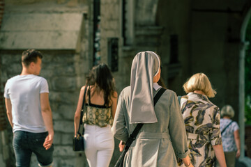 A Polish nun in a gray dress with a bag over her shoulder walks on a sunny day after a group of society people in casual clothes against the backdrop of the vintage stone walls of a medieval building