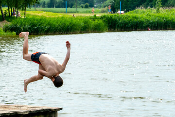 Teenagers jump into the water and swim in the lake on a hot summer day. Active recreation on an open pond. Children jump into the water and perform various acrobatic tricks.