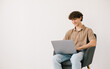 © anatoliycherkas - Happy young man using laptop while sitting in armchair against white wall, copy space. Millennial male communicating online, working or studying remotely on portable pc