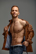 Portrait of a handsome male model posing shirtless in a brown leather jacket and jeans. Grey background. Fashionable autumn clothes. Men's youth fashion.