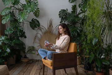 Wall Mural - Relaxed calm woman playing with spring toy for stress relief sit crossed legs in armchair. Female relieve stressful tension with stretchable spring has break from work at home office in indoor garden