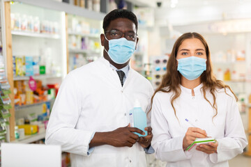 Portrait of two multinational pharmacists in protective masks, standing in the sales hall of a pharmacy with good during a ..pandemic and making important notes