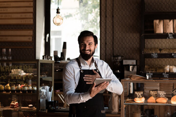 Happy smiling bearded man in apron (barista or small business owner) standing in a bakery cafe with tablet in hands