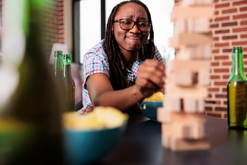 Wall Mural - Smiling woman sitting at table in living room while playing society games with friends. Joyful person pulling wooden block from wooden tower while sitting at home and enjoying fun leisure activity.