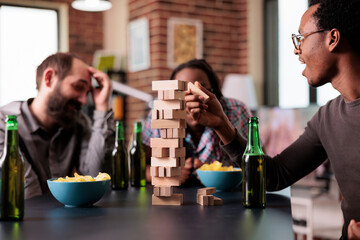 Wall Mural - Multiracial group of friends sitting at table in living room while playing together with wood blocks. Diverse people sitting at home while enjoying fun leisure activity having snacks and beverages.