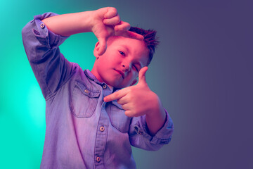 Portrait of little boy, child in jeans t-shirt posing isolated over green purple background in neon light. Looks delightful.