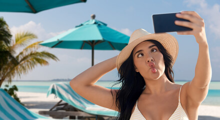 Wall Mural - travel, tourism and summer vacation concept - happy smiling young woman in bikini swimsuit and straw hat taking selfie with smartphone over tropical beach background in french polynesia