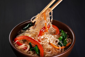 Rice glass noodles with vegetables bell peppers, carrots, spinach, sesame seeds and soy sauce. Chopsticks holding noodles.