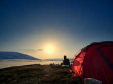 Person Sitting Next To A Red Tent In A Campsite Next To An Icy Lake Enjoying The Sunrise
