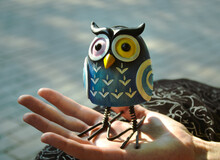 A Toy Of A Surprised Blue Owl With Multi-colored Eyes Close-up Stands On The Palm Of A Female Hand In The Light Of The Evening Sun