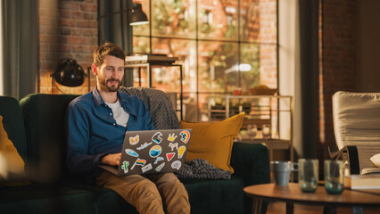 Poster - Portrait of a Handsome Man Sitting on Couch, Using Laptop Computer in Stylish Loft Apartment on a Sunny Day. Creative Male Smiling, Checking Social Media and Websites. Urban City View from Big Window.