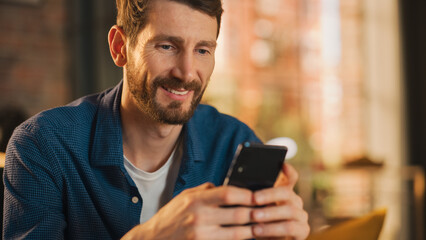 Fototapete - Portrait of a Handsome Man Sitting on Couch, Using Smartphone in Stylish Loft Apartment or Cafe. Creative Male Smiling, Checking Social Media and Typing Message. Urban City View from Big Window.