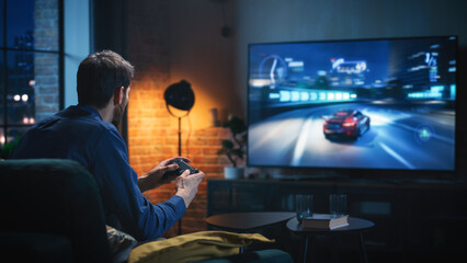 Wall Mural - Young Man Spending Time at Home, Sitting on a Couch in Stylish Loft Apartment and Playing Arcade Car Video Games on Console. Male Using Controller to Play Street Racing Drift Simulator.