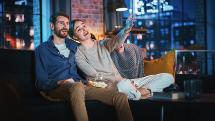 Wall Mural - Portrait of Beautiful Couple Spending Time at Home, Sitting on a Couch, Hugging and Watching Exciting TV Show in Their Stylish Loft Apartment. Man and Woman Streaming Comedy Movie and Have Good Time.