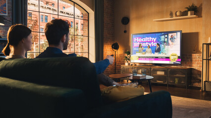 Aufkleber - Authentic Couple Spending Time at Home, Sitting on a Couch and Watching TV with Big Flat Screen Display in Their Stylish Loft Apartment. Man and Woman Streaming Reality Show or Infomercial.