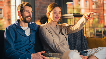 Wall Mural - Portrait of Young Couple Spending Time at Home, Sitting on a Sofa, Man Embracing His Girlfriend While Watching Exciting TV Show in Apartment. Man and Woman Streaming Movie and Get Surprised.