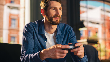 Portrait of Young Handsome Man Spending Time at Home, Sitting on Couch in Stylish Loft Apartment and Playing Video Games on Console. Successful Male Using Controller to Concentrate Fully on the Game.