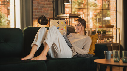 Poster - Young Beautiful Woman Lying Down on Couch, Use Laptop Computer in Stylish Loft Apartment on a Sunny Day. Creative Female Smiling, Checking Social Media. Urban City View from Big Window.