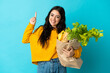 © luismolinero - Young woman holding a grocery shopping bag isolated on blue background showing and lifting a finger in sign of the best