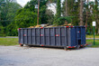 Side view of a long blue dumpster full of construction debris in front of green trees and shrubs